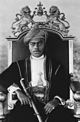 A black-and-white photograph of a man with a dark moustache wearing a turban and a dark jacket and sitting on a throne topped by two metal lions