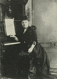 B&W photo of a middle-aged woman wearing a long dress, reclining at a piano