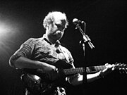 Schwarz-Weiß-Foto eines bärtigen Mannes, der auf der Bühne Gitarre spielt.