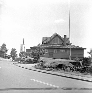 Fotografi från 1957 av Brukskapellet i Kallinge med Kallinge kyrka i bakgrunden. Foto av Armin Tuulse genom Riksantikvarieämbetet.