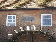 Bâtiment en briques avec toit de tuiles et deux fenêtres carrées, au-dessous duquel se trouve une roue à eau.  Entre les fenêtres, il y a une plaque en bois, qui se lit "Cobham Mill restauré 1993 par Cobham Mill Preservation Trust et la National Rivers Authority".