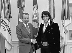 Um Presley com corte de carneiro, vestindo uma longa jaqueta de veludo e uma fivela gigante como a de um cinturão de campeão de boxe, aperta a mão de um homem careca de terno e gravata.  Eles estão enfrentando a câmera e sorrindo.  Cinco bandeiras estão penduradas em mastros logo atrás delas.