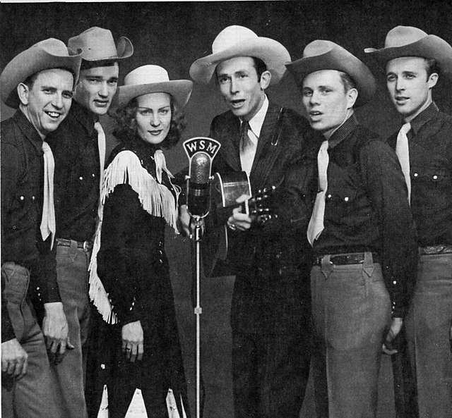 A group of six men and one woman wearing cowboy hats, standing around a microphone.  The man third from right is holding a guitar.