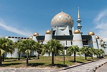 Fotografia di una moschea bianca e dorata, la sua grande cupola, il suo alto minareto e le poche palme che la circondano.