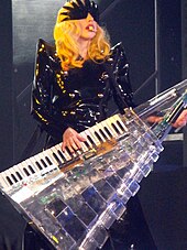 Profile of a young blond woman. Her hair falls in waves up to her shoulders. She wears a hairy dress with a shiny cloth wrapped around her.