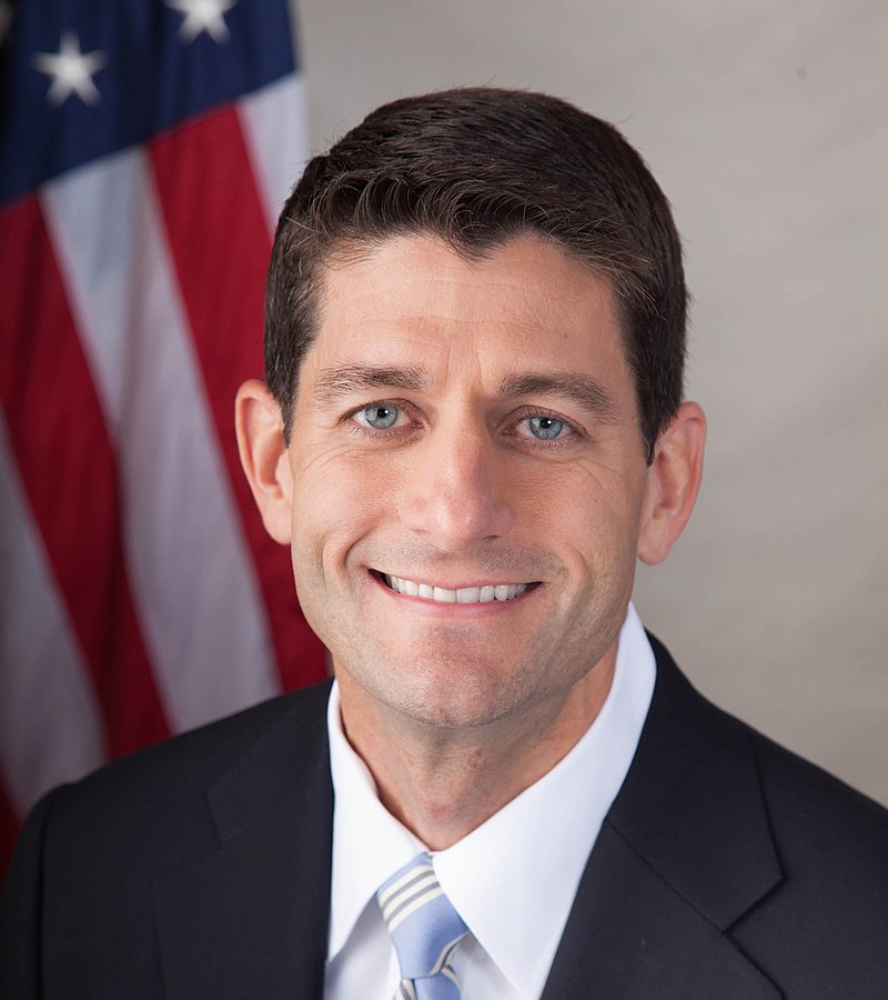 A portrait shot of Paul Ryan, looking straight ahead. He has short brown hair, and is wearing a dark navy blazer with a red and blue striped tie over a light blue collared shirt. In the background is the American flag.