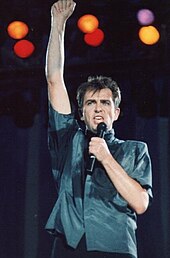A man wearing a blue-shade shirt performing on a microphone on one hand while raising the other