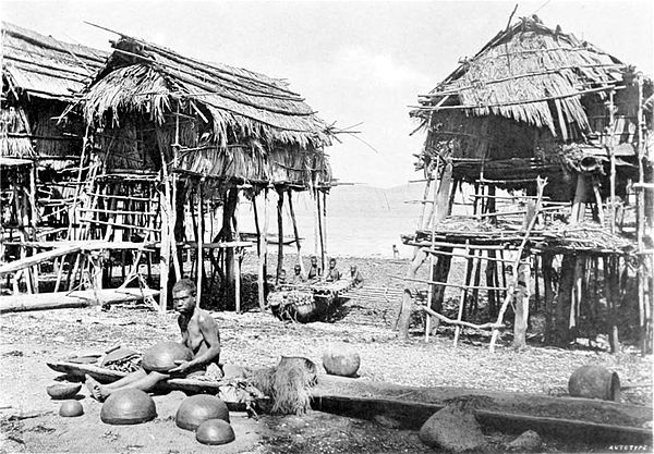 Black and white photograph of a woman making pots on a beach, with three huts-on-stilts in the background.