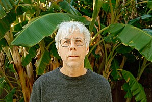 John Vanderslice wearing a grey sweater, standing in front of a banana palm in Los Angeles