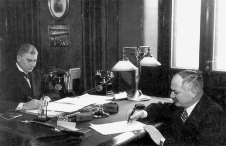 Two men sign papers at opposite sides of a table in a small office.