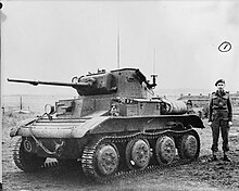 Un tanque pequeño y achaparrado se encuentra en un campo, con las vías de la izquierda hacia la cámara.  El cañón de su arma se ha alargado con una extensión de metal.  Un hombre de uniforme con boina negra está junto a él.  Al fondo, se ven campos a la izquierda y varias casas a la derecha.