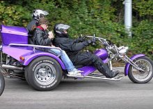Photo couleur d'un tricycle motorisé, avec un conduxteur casqué et deux passagers.