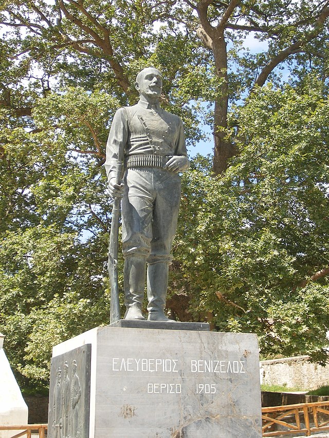 Statue en bronze d'un homme en armes sur un piédestal gris, dans les arbres.