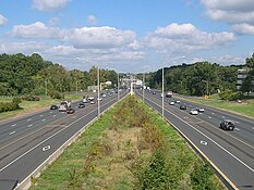 Both sides of a highway with a grass plot in the middle of the roads. Street lamps surround the middle, and several cars are on the roads. The roads have an HOV diamond on them.