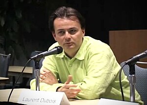 A man sits behind a table at a microphone. In front of him a placard reads Laurent Dubois.