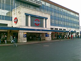 Entrée de la gare sous la forme d'une boîte revêtue de pierre blanche reposant sur deux blocs de pierre importants et larges.  La façade avant de la boîte contient un grand logo du métro de Londres (anneau rouge avec une barre horizontale bleue au centre contenant le mot "UNDERGROUND") au centre.  En retrait derrière l'entrée et de part et d'autre, s'élève un immeuble de bureaux de quatre étages au bardage bleu.