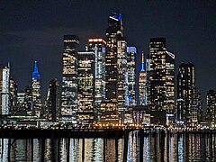 Teleaufnahme bei Nacht über den an der Oberfläche sanft gekräuselten Hudson River hinweg. Viele bunte, vor allem blaue, Akzentlichter von den Hochhäusern Hudson Yards im Mittelpunkt und von One Vanderbilt und Empire State Building im Hintergrund.