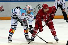 Fotografia a colori di due giocatori di hockey fianco a fianco, bastoni appoggiati sul ghiaccio davanti a loro, trasversalmente.