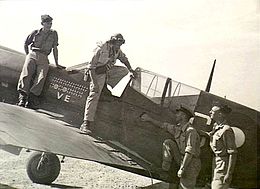 Piloot met veiligheidsbril die tevoorschijn komt uit de cockpit van een eenmotorige eendekker met de letters "VE" prominent op de romp, in gezelschap van drie andere mannen