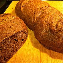 Due pagnotte di pane, una con un lato tagliato a sinistra e l'altra intera, di colore marrone su un tavolo di legno chiaro.