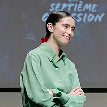 Sara Montpetit, a white young woman with black long hair which is tied into a ponytail with red clamp, smiling and looking off to the side with her arms folded wearing a lime green blouse