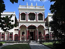 Through gardens and a fence, a two storey building can be seen. The front door is protected by Chinese guardian lions. The facade has Victorian columns and features.