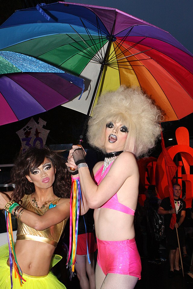 Twee dragqueens in korte broek en croptop met ieder een regenboogparaplu