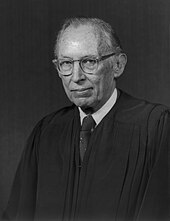 retrato fotográfico monocromático de um homem branco no final da meia-idade em vestes judiciais.  Ele usa óculos e tem cabelos ralos.