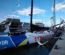 Vue du bateau de trois-quarts avant. On voit le mât, les outriggers, le foil bâbord, le rouf, mais on ne voit pas l'avant.