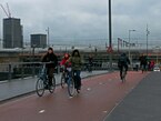 Amsterdam, 2023 - People are biking in gray, rainy weather over the bridge Odebrug, to cross the Prins Hendrikkade direction Gelderse Kade. On the background the train track at the East side of Central Station - free download street photography by Fons Heijnsbroek