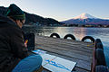 An elderly Japanese man sittiong on a small wooden pier drawing Mount Fuji as seen at sunrise across lake Kawaguchi, with Fujikawaguchiko town in the foreground. Honshu Island. Japan