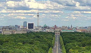 Farbfotografie von Berlin in der Vogelperspektive. Im Vordergrund stehen bewaldete Flächen vom Park Tiergarten und die Straße des 17. Juni führt zum Brandenburger Tor. Von links nach rechts sind das Reichstagsgebäude, der Fernsehturm, der Berliner Dom, das Rote Rathaus und das Berliner Schloss