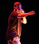 An Afro-American man wearing a black shirt, on stage while holding a microphone.