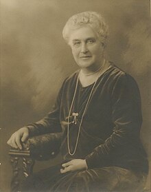 An older white woman with white hair, seated in a carved wooden chair, wearing a loose-fitting dark velvet dress with long sleeves