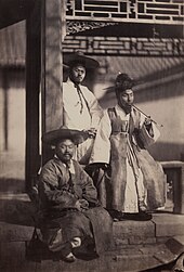 Photographie en noir et blanc montrant trois hommes au pied d’un bâtiment, celui de gauche est assis sur un muret et porte un couvre-chef à large bord, derrière lui le second est debout et appuyé contre un poteau ; assis entre les deux, le troisième fume une pipe allongée.