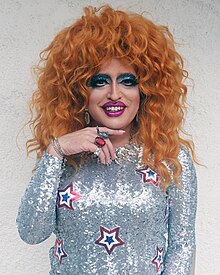 Drag queen Lil Miss Hot Mess smiles at the camera, posing with her chin resting on her finger. She has light red curly hair, and ironically wears a silver sequin dress with red-white-and-blue sequin stars.