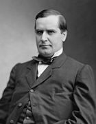 A terse, professional-looking man reclines in a chair in this black-and-white photograph. He has a prominent nose and dimple in his chin. His dark jacket has only the top button fastened, and appears to be under some strain.