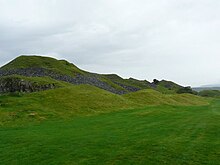 Photo d'un paysage vallonné recouvert d'herbe sous un ciel nuageux. On distingue les ruines de murs de pierre grise