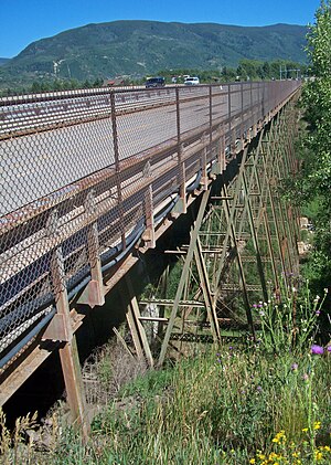 A metallic bridge with wide steelwork supports over a grassy gorge with tree-covered mountains in the background, seen from its right