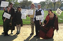 Photo de groupe de six personnes en costume, portant des écriteaux avec des slogans en rapport avec la série