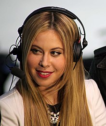 Caucasian woman in her late 30s with long and straight blonde hair, wearing a white jacket and black headset