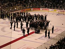 Члены хоккейной команды вместе поднимают трофей на праздновании, в то время как многочисленные высокопоставленные лица и представители СМИ окружают их.