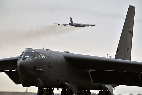 A B-52H Stratofortress of the 5th Bomb Wing sits on the flight-line as another B-52H takes off from Minot AFB.