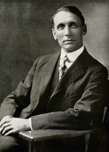 Black and white photograph of a man in an overcoat, sitting in a chair visible from the waist up. He is clean shaven, has his hair combed to the side. He wears a calm expression and is looking to the left towards the camera.