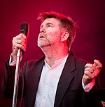 A man holding a microphone and looking up, with a red stage light beaming down
