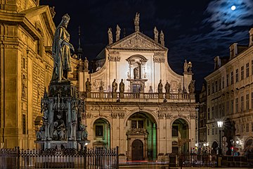 La façade d'une église baroque, de nuit, au clair de lune, entourée par plusieurs bâtiments, une statue d'un homme au premier plan à gauche.