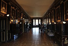 A photograph of a long dark room with high plain ceilings and gold-framed portraits lining the walls