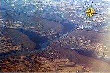 Oblique air photo, facing southwest, of the Potomac River flowing through water gaps in the Blue Ridge Mountains. Virginia on the left, Maryland on the right, West Virginia in upper right, including Harpers Ferry (partially obscured by Maryland Heights of Elk Ridge Mountain) at the confluence of the Potomac and Shenandoah Rivers. Potomac River Water Gaps Harpers Ferry w compass and pointer.jpg