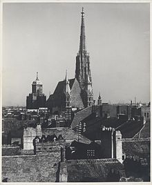 Fotografie alb-negru a unei catedrale care iese dintr-o mare de acoperișuri, turnul ei ieșind în evidență pe un cer senin