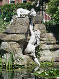 Colour photograph of two nude female statues, one on top of a rock helping another climb up it